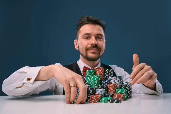 Hombre en chaleco negro y camisa sentado en la mesa blanca con montones de fichas en él, posando sobre fondo de estudio azul. Apuestas, póquer, casino. Primer plano . —  Fotos de Stock