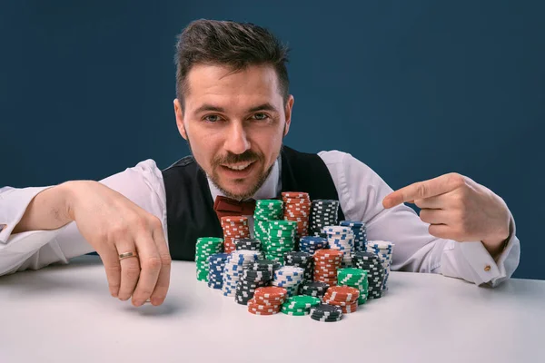 Чоловік в чорному жилеті і сорочці, сидячи за білим столом зі стеками чіпсів на ньому, позує на синьому студійному фоні. Азартні ігри, покер, казино. Крупним планом . — стокове фото