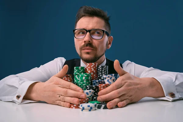 Хлопець в окулярах, чорний жилет і сорочка, сидячи за білим столом зі стосами чіпсів на ньому, позує на синьому фоні. Азартні ігри, покер, казино. Крупним планом . — стокове фото
