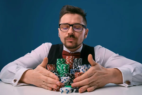 Чоловік в окулярах, чорний жилет і сорочка, сидячи за білим столом зі стосами чіпсів на ньому, позує на синьому фоні. Азартні ігри, покер, казино. Крупним планом . — стокове фото