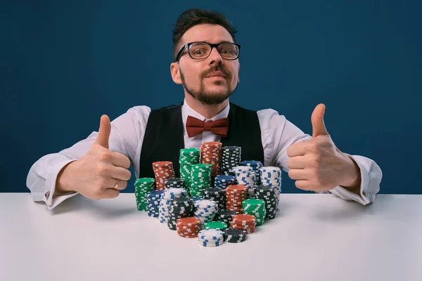 Гравець в окулярах, чорний жилет і сорочка, сидячи за білим столом зі стосами чіпсів на ньому, позує на синьому фоні. Покер, казино. Крупним планом . — стокове фото
