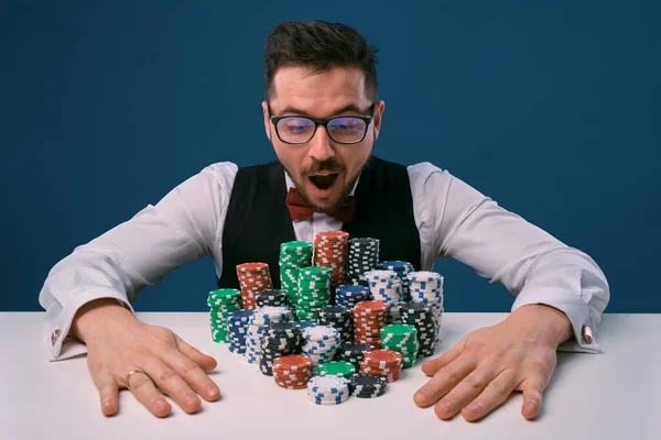 Tipo con gafas, chaleco negro y camisa sentado en la mesa blanca con montones de fichas en ella, posando sobre fondo azul. Apuestas, póquer, casino. Primer plano . —  Fotos de Stock