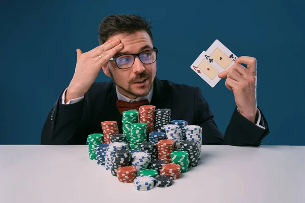 Man in glasses, black suit sitting at white table with stacks of chips, holding two playing cards, posing on blue background. Poker, casino. Close-up. — Stockfoto