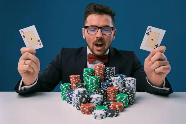 Fellow in glasses, black suit sitting at table with stacks of chips, holding two playing cards, posing on blue background. Poker, casino. Close-up. — 图库照片