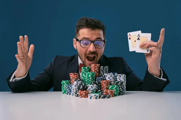 Player in glasses, black suit sitting at table with stacks of chips, holding two playing cards, posing on blue background. Poker, casino. Close-up. — Stockfoto