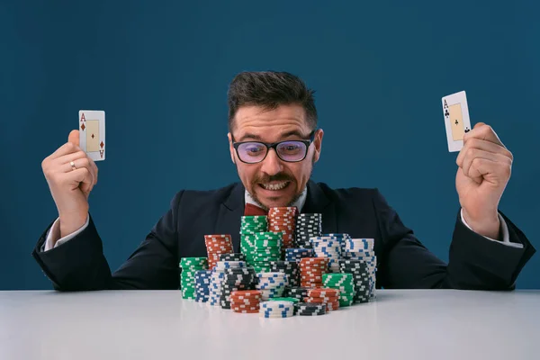 Fellow in glasses, black suit sitting at table with stacks of chips, holding two playing cards, posing on blue background. Poker, casino. Close-up. — 图库照片