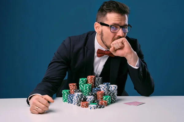 Male in glasses, black suit looking upset, sitting at white table with colored stacks of chips on it, posing on blue background. Poker, casino — 图库照片