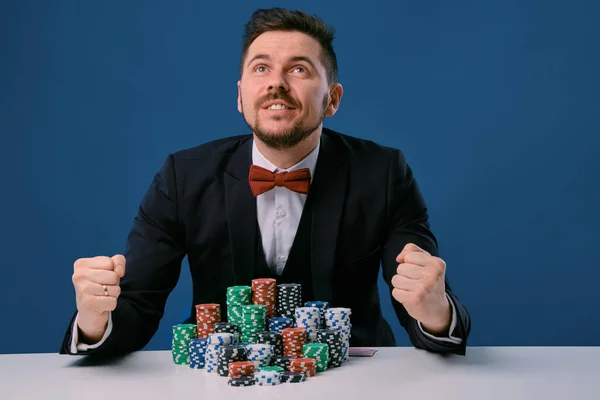 Чоловік у чорному костюмі сидить за білим столом з кольоровими стеками чіпсів на ньому, позуючи на синьому студійному фоні. Азартні ігри, покер, казино. Крупним планом . — стокове фото