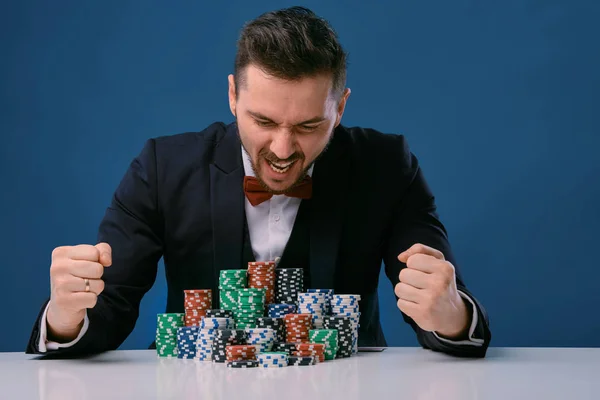 Man in black suit is sitting at white table with colored stacks of chips on it, posing on blue studio background. Gambling, poker, casino. Close-up. — Stockfoto