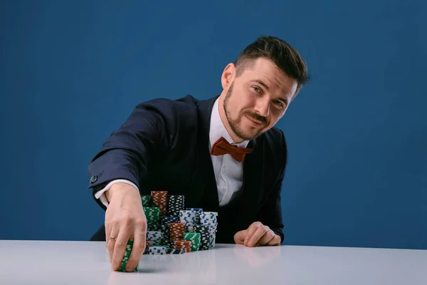 Man in black suit is sitting at white table with colored stacks of chips on it, posing on blue studio background. Gambling, poker, casino. Close-up. — ストック写真