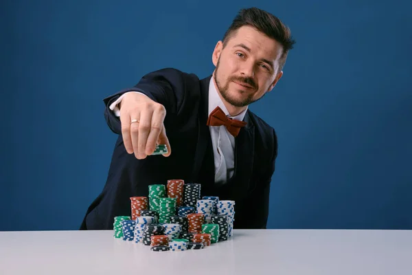 Чоловік у чорному костюмі сидить за білим столом з кольоровими стеками чіпсів на ньому, позуючи на синьому студійному фоні. Азартні ігри, покер, казино. Крупним планом . — стокове фото