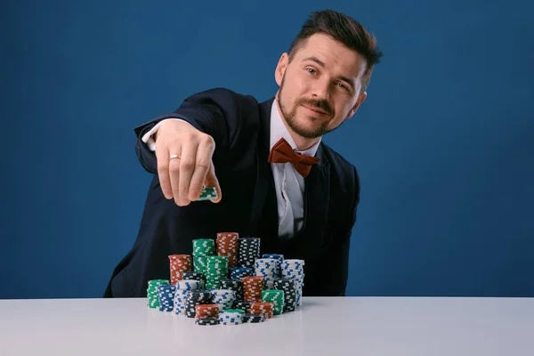 Man in black suit is sitting at white table with colored stacks of chips on it, posing on blue studio background. Gambling, poker, casino. Close-up. — ストック写真