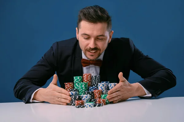 Uomo in abito nero è seduto al tavolo bianco con pile colorate di patatine su di esso, in posa su sfondo blu studio. Gioco d'azzardo, poker, casinò. Primo piano . — Foto Stock