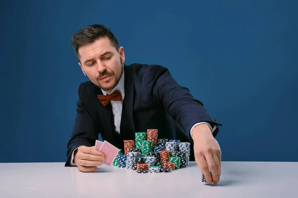 Man in black suit is sitting at white table with stacks of chips, holding two playing cards, posing on blue background. Poker, casino. Close-up. — Stockfoto