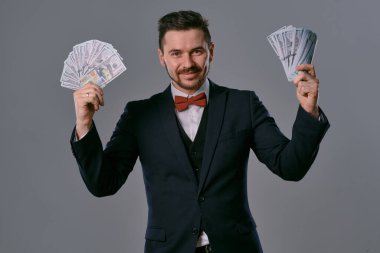 Man in black suit and red bow-tie is showing two fans of hundred dollar bills, posing on gray studio background. Gambling, poker, casino. Close-up.