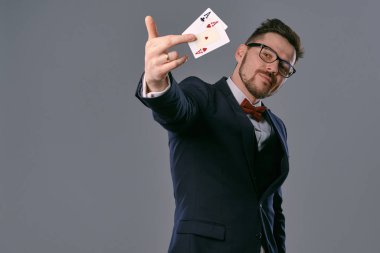 Man in black classic suit, red bow-tie, glases is showing two playing cards, posing on gray studio background. Gambling, poker, casino. Close-up.