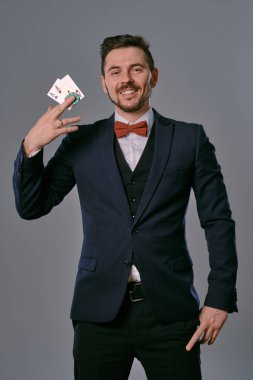 Man in black classic suit and red bow-tie showing two playing cards and chips, posing on gray studio background. Gambling, poker, casino. Close-up.