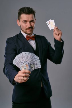 Man in black classic suit and red bow-tie showing two playing cards and cash, posing on gray studio background. Gambling, poker, casino. Close-up.