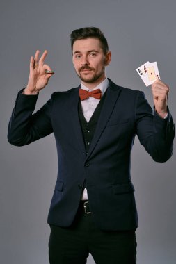 Man in black classic suit and red bow-tie showing two playing cards and chips, posing on gray studio background. Gambling, poker, casino. Close-up.