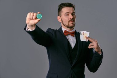 Man in black classic suit and red bow-tie showing two playing cards and chip, posing on gray studio background. Gambling, poker, casino. Close-up.