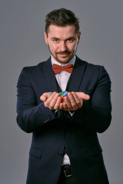 Man in black classic suit and red bow-tie showing some colored chips, posing against gray studio background. Gambling, poker, casino. Close-up.