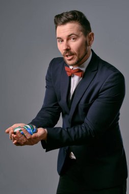 Man in black classic suit and red bow-tie showing some colored chips, posing against gray studio background. Gambling, poker, casino. Close-up.