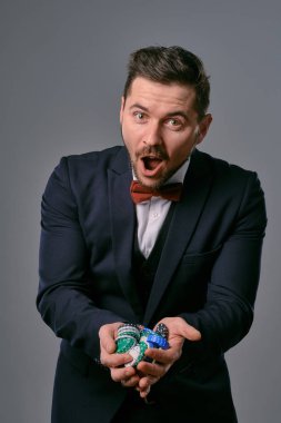 Man in black classic suit and red bow-tie showing some colored chips, posing against gray studio background. Gambling, poker, casino. Close-up.
