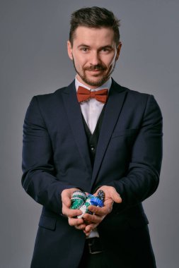 Man in black classic suit and red bow-tie showing some colored chips, posing against gray studio background. Gambling, poker, casino. Close-up.