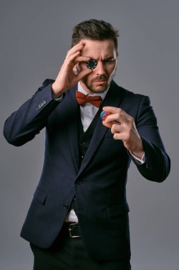 Man in black classic suit and red bow-tie showing some colored chips, posing against gray studio background. Gambling, poker, casino. Close-up.