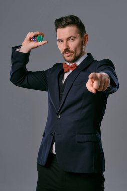 Man in black classic suit and red bow-tie showing some colored chips, posing against gray studio background. Gambling, poker, casino. Close-up.