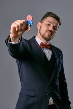 Man in black classic suit and red bow-tie showing some colored chips, posing against gray studio background. Gambling, poker, casino. Close-up.