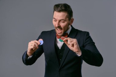 Man in black classic suit and red bow-tie showing some colored chips, posing against gray studio background. Gambling, poker, casino. Close-up.