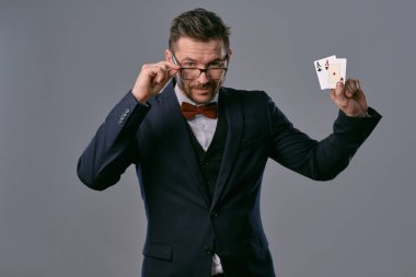 Man in black classic suit, red bow-tie, glases is showing two playing cards, posing on gray studio background. Gambling, poker, casino. Close-up.