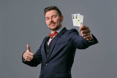 Man in black classic suit and red bow-tie showing two playing cards while posing against gray studio background. Gambling, poker, casino. Close-up.