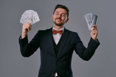 Man in black suit and red bow-tie is showing two fans of hundred dollar bills, posing on gray studio background. Gambling, poker, casino. Close-up.