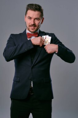 Man in black classic suit and red bow-tie showing two playing cards while posing against gray studio background. Gambling, poker, casino. Close-up.