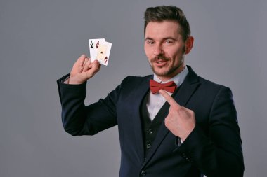 Man in black classic suit and red bow-tie showing two playing cards while posing against gray studio background. Gambling, poker, casino. Close-up.
