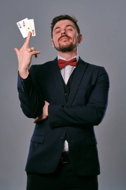 Man in black classic suit and red bow-tie showing two playing cards while posing against gray studio background. Gambling, poker, casino. Close-up.