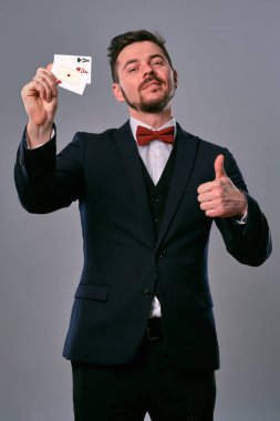 Man in black classic suit and red bow-tie showing two playing cards while posing against gray studio background. Gambling, poker, casino. Close-up.