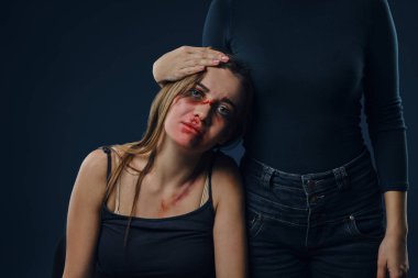 Female in black blouse has put her hand on head of a girl. Victim with bruises on face sitting nearby. Blue background. Domestic violence. Close-up.