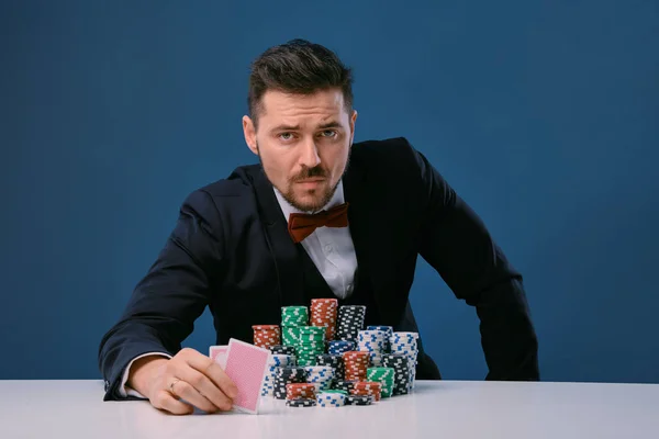 Man in black suit is sitting at white table with stacks of chips, holding two playing cards, posing on blue background. Poker, casino. Close-up. — Stockfoto