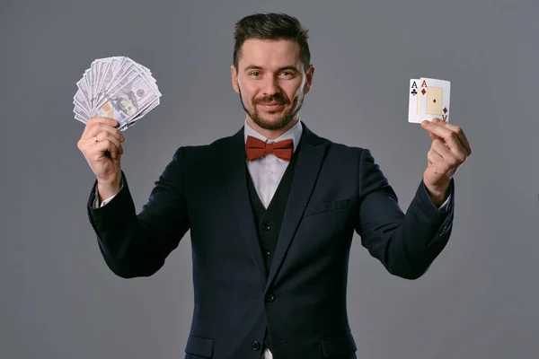 Man in black suit and red bow-tie is showing two fans of hundred dollar bills, posing on gray studio background. Gambling, poker, casino. Close-up.