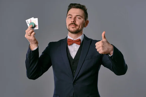 Man in black classic suit and red bow-tie showing two playing cards and chips, posing on gray studio background. Gambling, poker, casino. Close-up. — Φωτογραφία Αρχείου