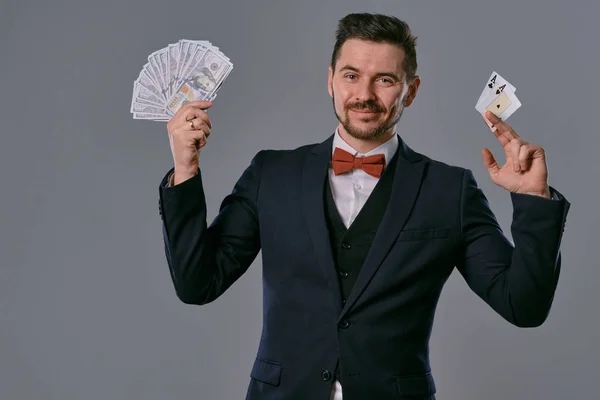 Hombre de traje clásico negro y pajarita roja mostrando dos cartas y dinero en efectivo, posando sobre fondo gris estudio. Apuestas, póquer, casino. Primer plano . —  Fotos de Stock