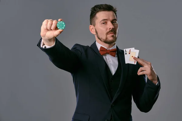 Man in black classic suit and red bow-tie showing two playing cards and chip, posing on gray studio background. Gambling, poker, casino. Close-up. — ストック写真