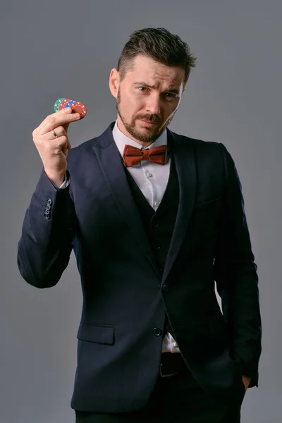Man in black classic suit and red bow-tie showing some colored chips, posing against gray studio background. Gambling, poker, casino. Close-up. — ストック写真