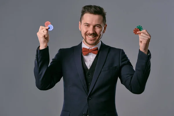 Man in black classic suit and red bow-tie showing some colored chips, posing against gray studio background. Gambling, poker, casino. Close-up. — Stockfoto