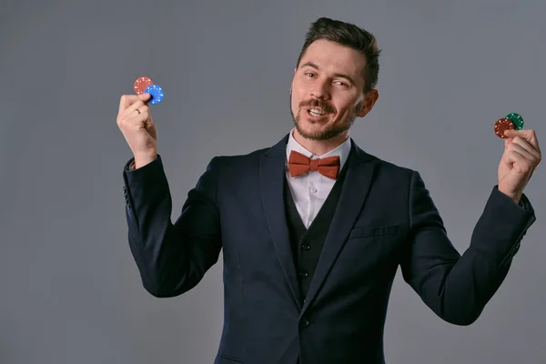 Man in black classic suit and red bow-tie showing some colored chips, posing against gray studio background. Gambling, poker, casino. Close-up. — ストック写真