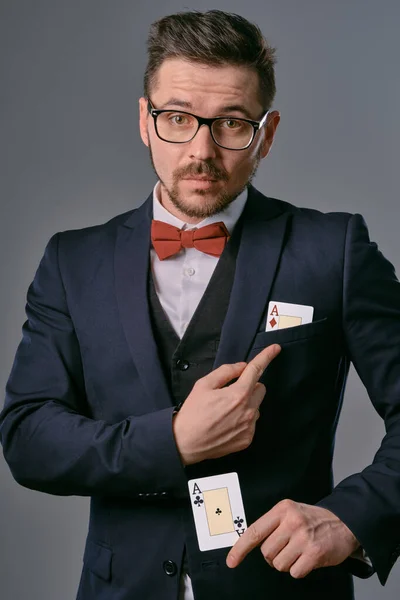 Man in black classic suit, red bow-tie, glases is showing two playing cards, posing on gray studio background. Gambling, poker, casino. Close-up. — Stockfoto