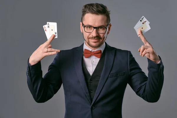 Man in black classic suit, red bow-tie, glases is showing four playing cards, posing on gray studio background. Gambling, poker, casino. Close-up. — Stockfoto